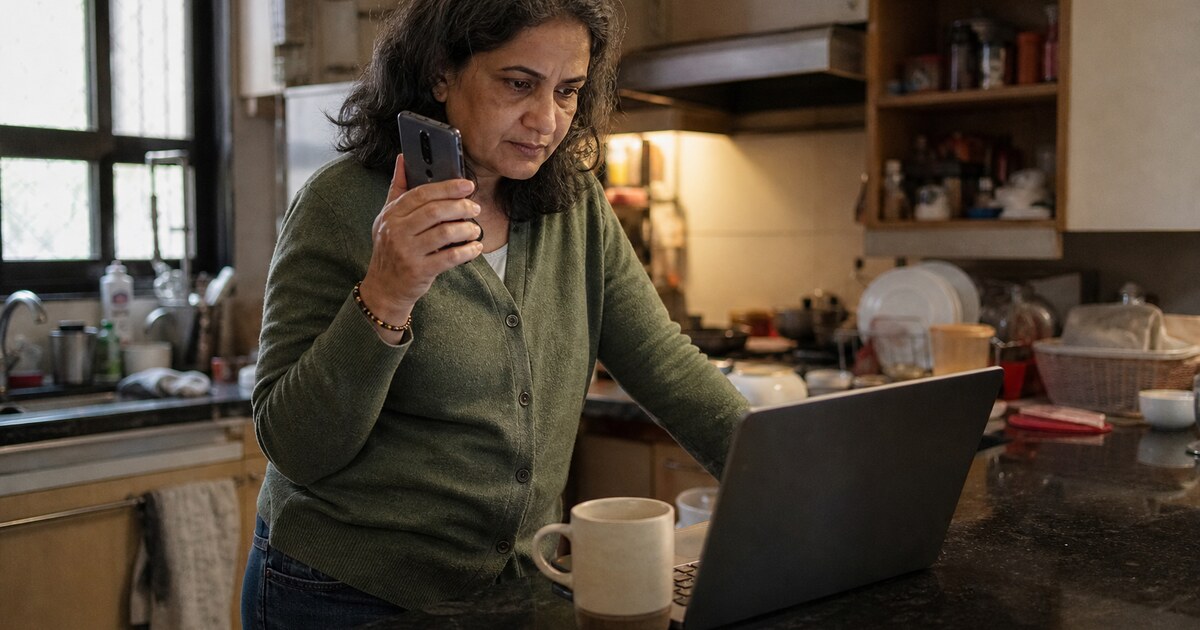 A South Asian woman in her late forties with shoulder-length greying hair, wearing a faded green cardigan and jeans, stands at a cluttered…