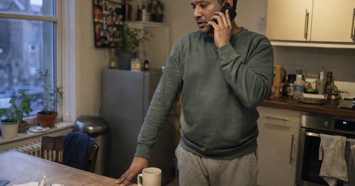 A 34-year-old South Asian man with short curly hair, wearing a faded green sweatshirt and grey joggers, stands beside a cluttered kitchen…